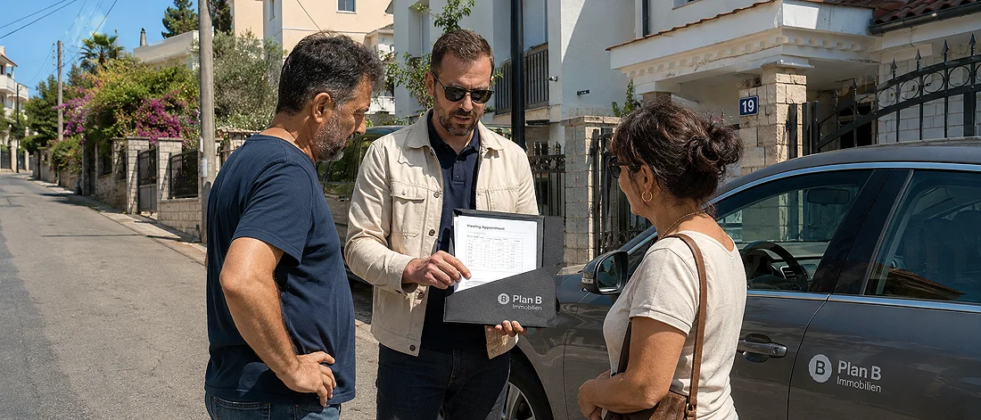 Ein Mann, der eine Mappe in der Hand hält, zeigt einem Mann und einer Frau, die neben einem geparkten Auto mit der Aufschrift „Plan B Immobilien Montenegro“ in einer Wohnstraße stehen, einige Unterlagen.