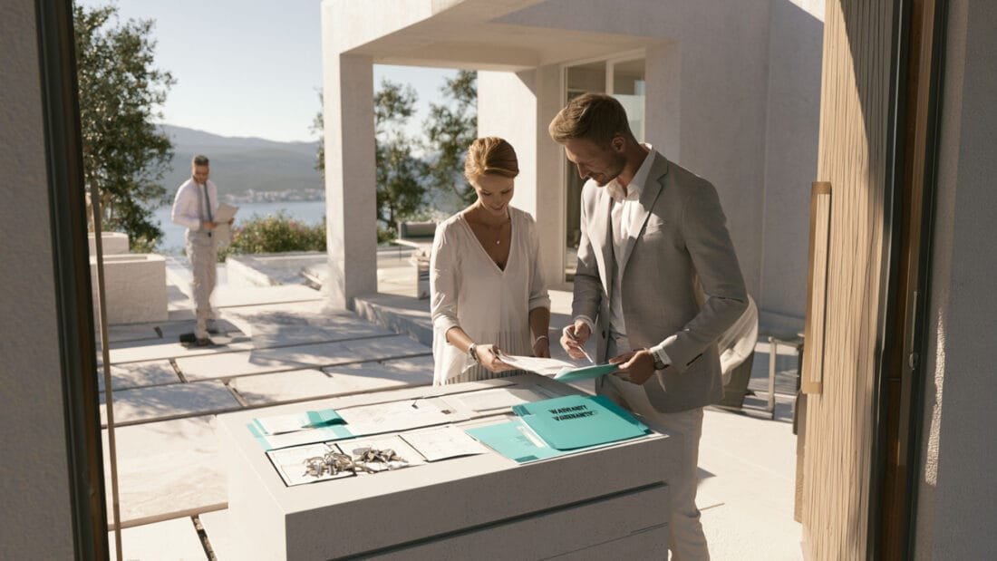 Plan B Immobilien Montenegro Ulcinj - Ein Mann und eine Frau in formeller Kleidung sehen sich draußen an einem Tisch Dokumente an; dabei sind Hausschlüssel und Ordner zu sehen. Im Hintergrund ist ein weiterer Mann in weißer Kleidung zu sehen, der in der Nähe eines modernen Hauses spaziert, von dem aus man einen Blick auf das Wasser und die Berge hat.
